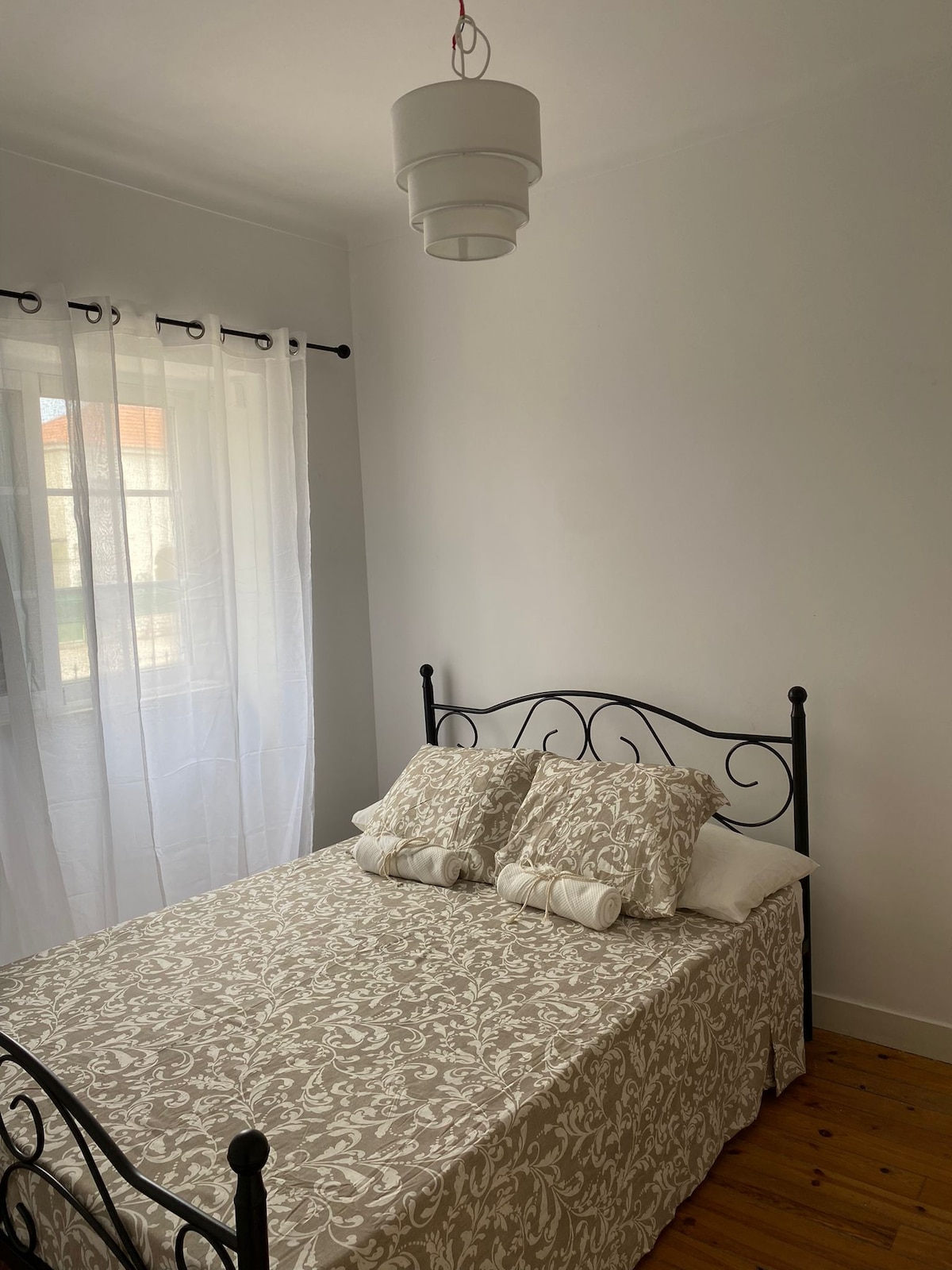 A sleeping area features a decorative bed frame with a patterned bedspread and neatly arranged pillows. Soft light filters through sheer curtains covering the window, enhancing the simplicity of the space with light-colored walls and wooden flooring.
