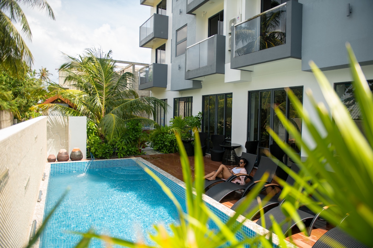 A serene pool area is surrounded by lush greenery and sun loungers. Balconies from the upper floors provide views of the outdoor space, where sunlight reflects off the clear water of the swimming pool, creating a relaxing atmosphere.