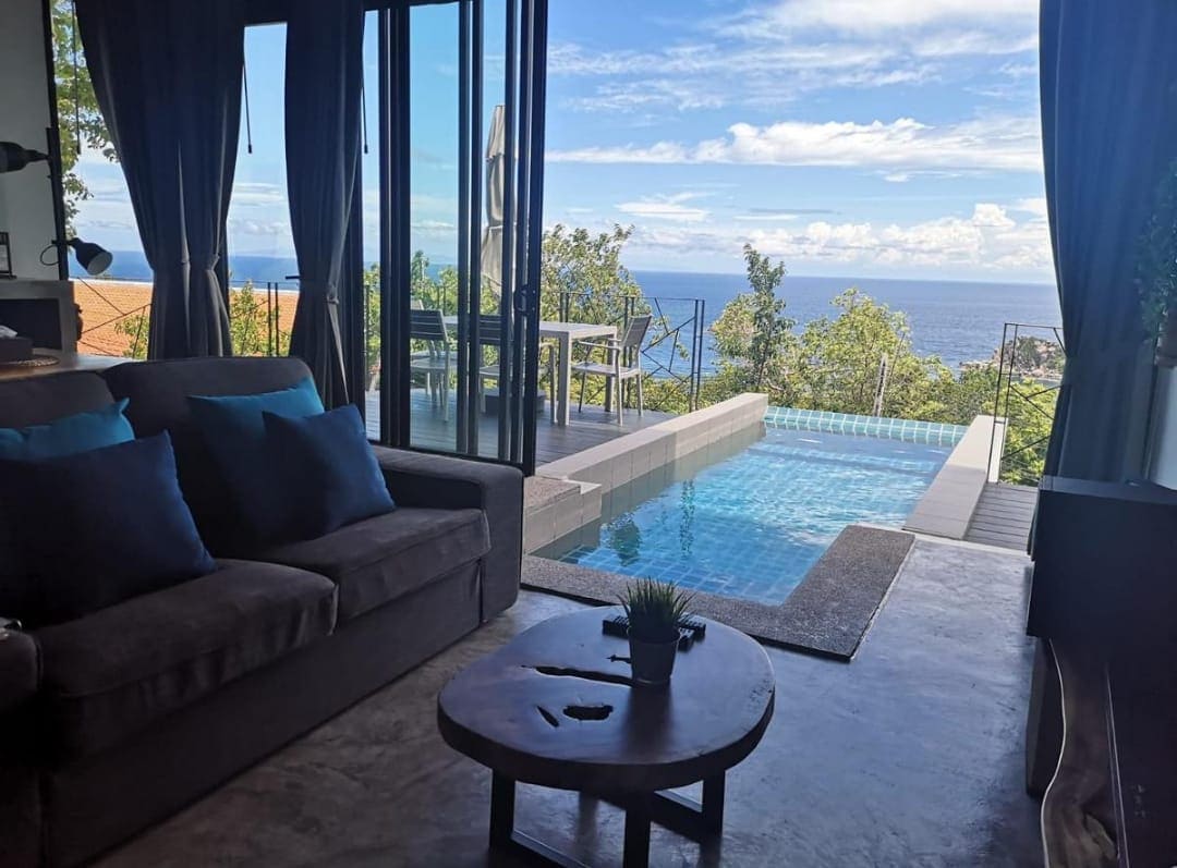 A comfortable living area is visible, featuring a dark sofa adorned with blue cushions. Large glass doors open to reveal a private pool and sweeping ocean views, framed by lush greenery. Sunlight brightens the modern space, creating a serene atmosphere.