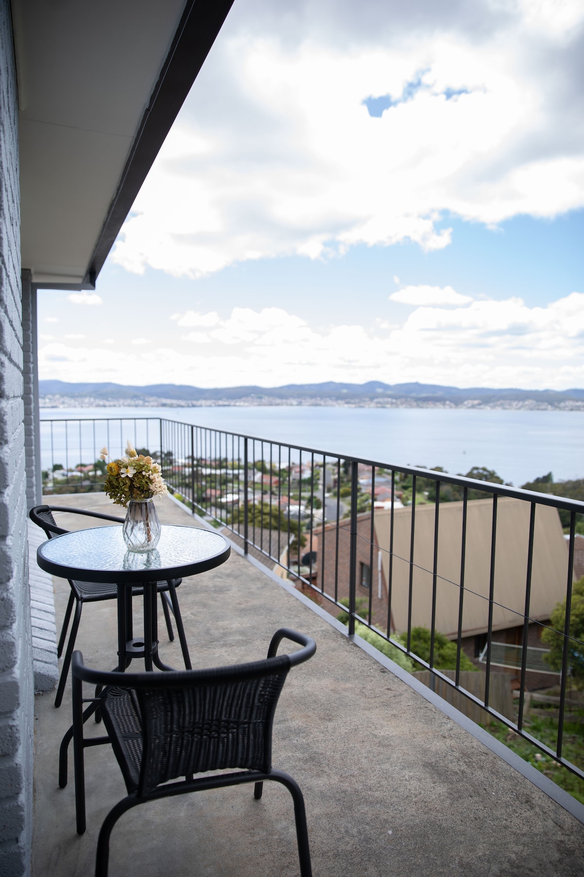 An outdoor balcony with a round black table and two matching chairs overlooks a scenic ocean view. A small vase with fresh flowers is centered on the table. The sky is partly cloudy, enhancing the serene ambiance of the surrounding landscape.