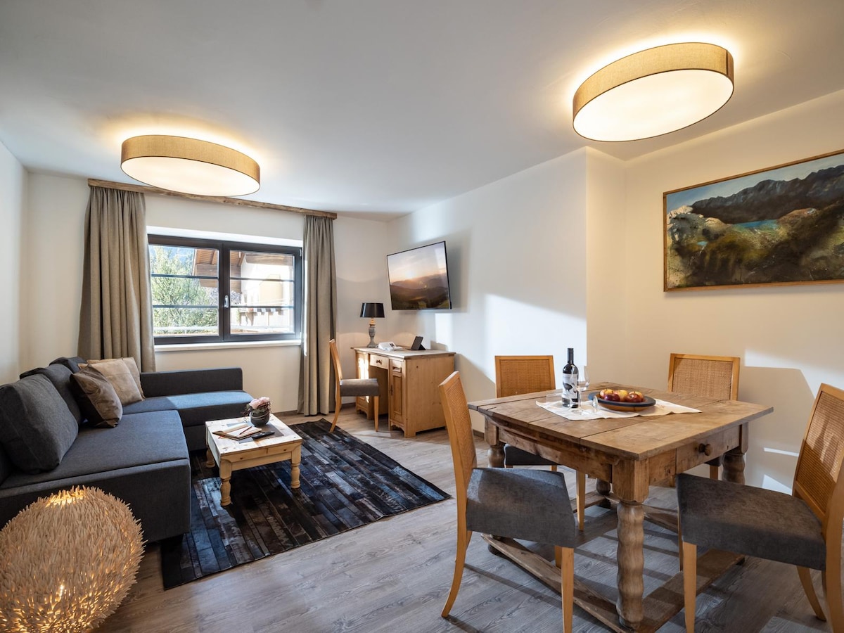 A bright living area is furnished with a gray sofa and a wooden dining table. Natural light enters through a window, illuminating the space. A television is mounted on the wall, and a piece of artwork adds color to the room. Soft lighting is provided by two ceiling fixtures.