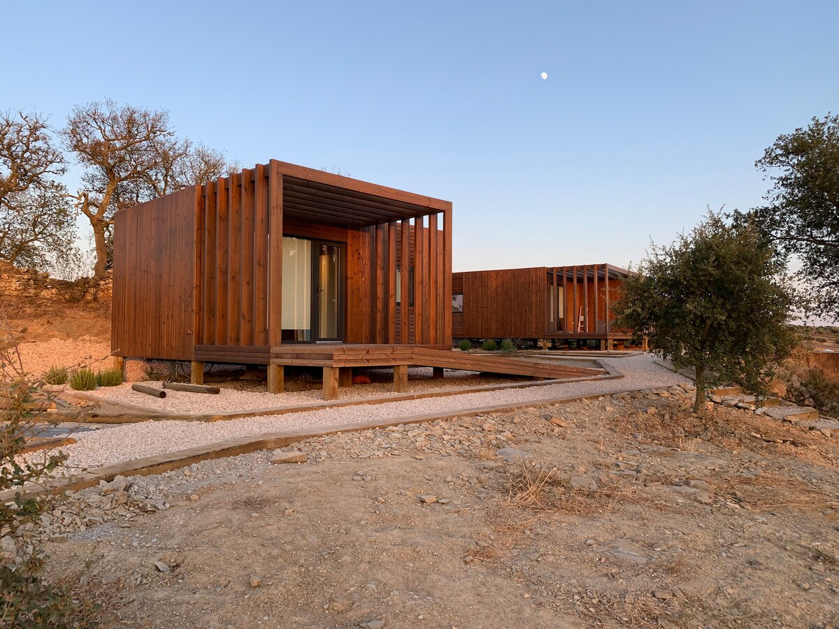 Two unique wooden structures are positioned on a gravel pathway surrounded by a natural landscape. The angular design features horizontal slats, allowing glimpses of the interiors. Soft colors of the sunset paint the sky, enhancing the rustic charm of this tranquil setting.
