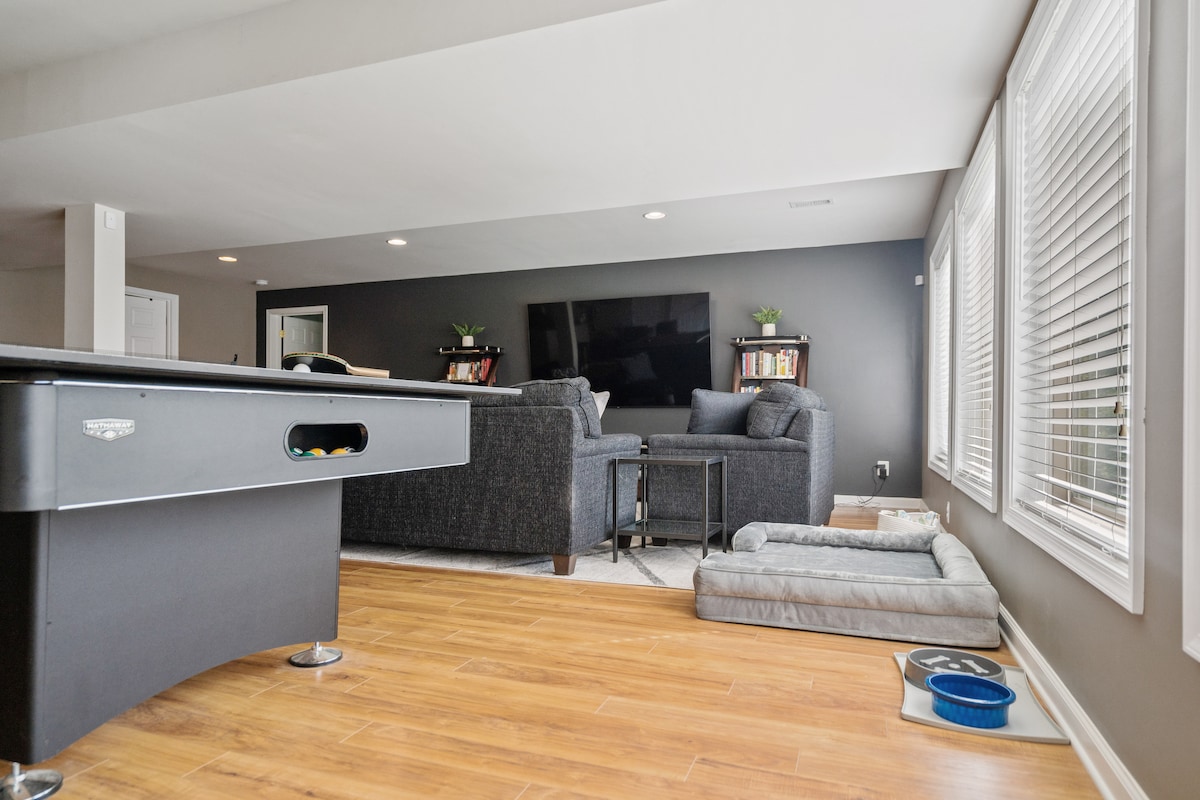 An inviting indoor space includes a billiards table positioned beside a dark grey couch and armchair. Natural light floods through large windows dressed with white blinds. A small table stands in front of the seating area, while a dog bed and food dishes are placed on the floor.