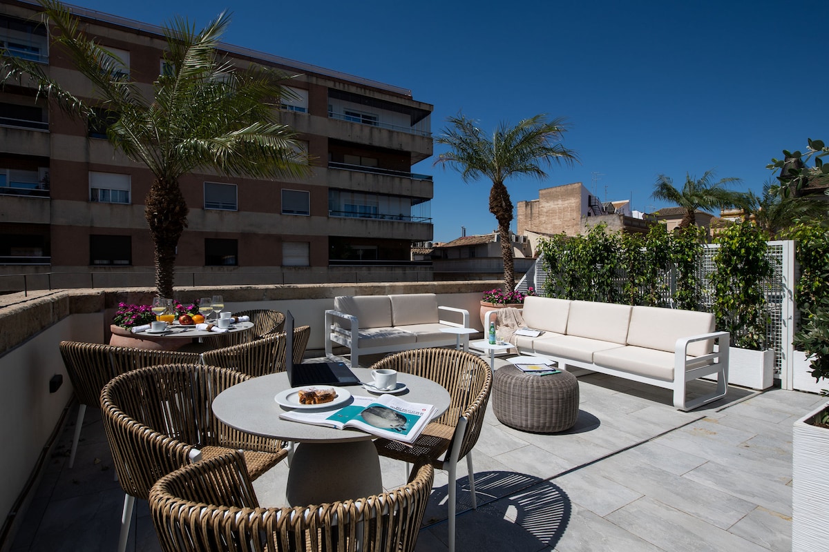 A spacious terrace is depicted, furnished with circular tables and comfortable seating arrangements. Lush greenery and palm trees provide a tropical feel. Warm sunlight brightens the area, inviting relaxation and outdoor dining experiences.