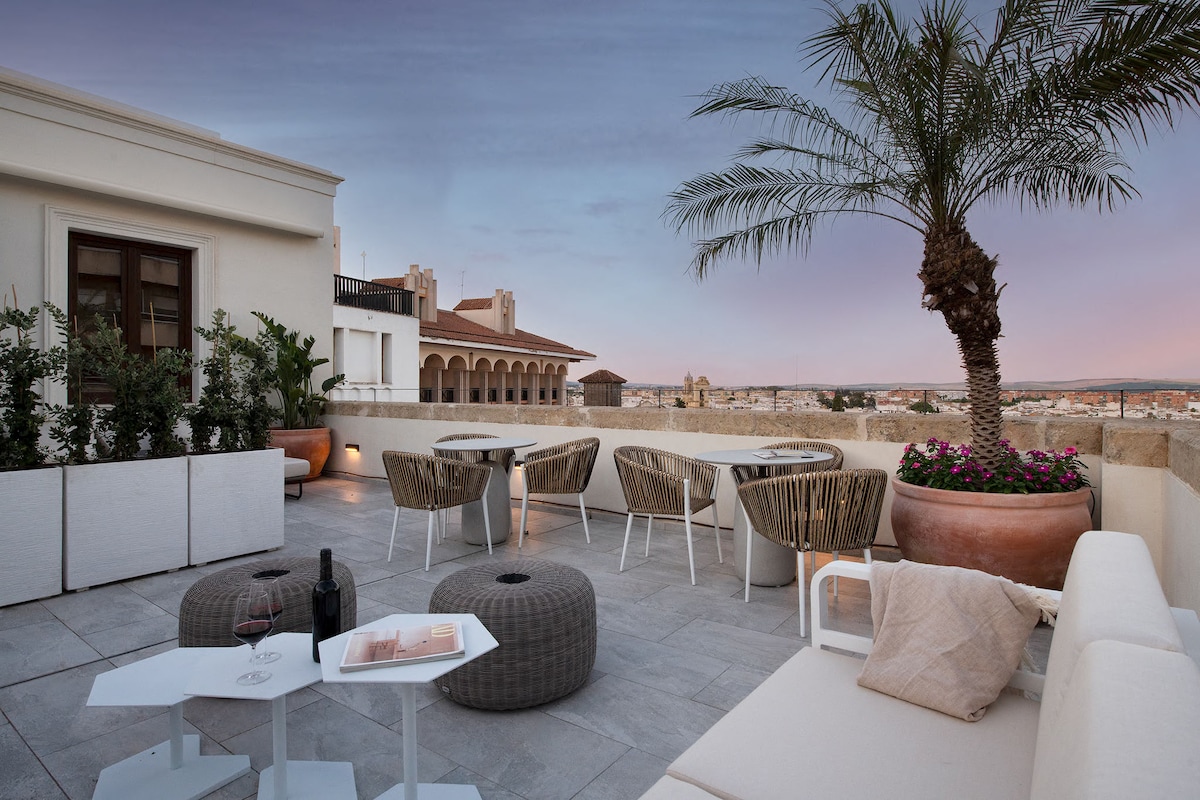 A rooftop terrace features modern outdoor furniture, including lounge chairs and a small table. Potted plants and a palm tree enhance the space. Soft evening light illuminates the area, providing a serene setting with views of the surrounding city landscape.