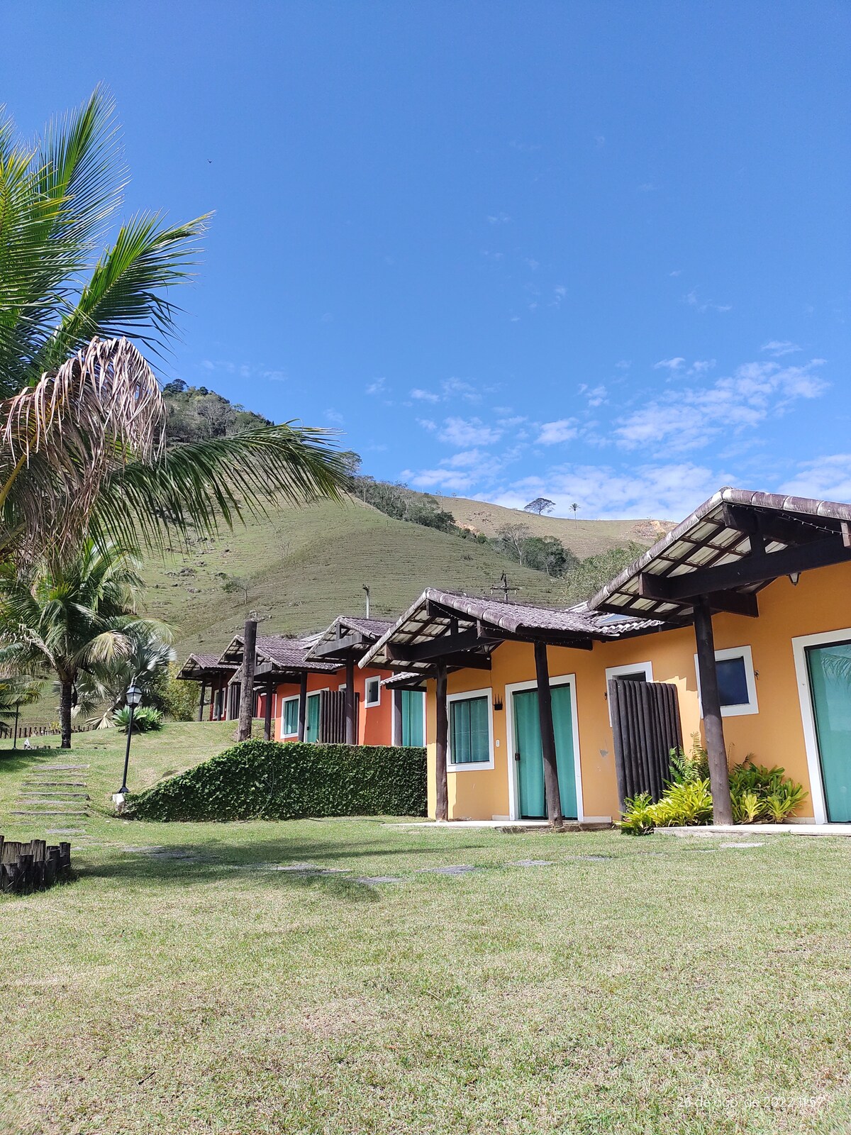 A row of chalets is positioned on a gentle hillside, featuring colorful exteriors surrounded by lush green grass. Palm trees add a tropical touch alongside the serene landscape, with rolling hills visible in the background under a clear blue sky.