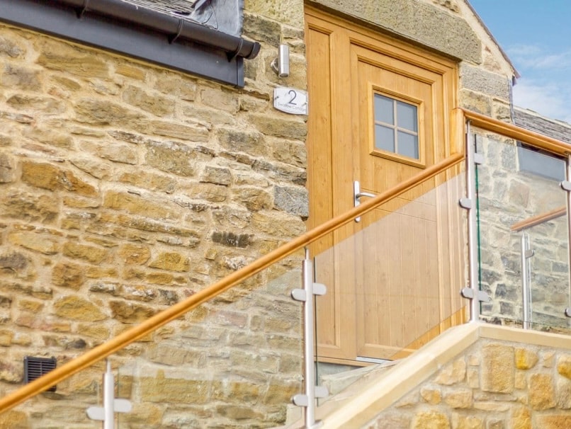 The entrance of the cottage features a wooden door with a small window, surrounded by rustic stone walls. A set of 15 open tread steps leads up to the door, accented by a glass and wooden railing.