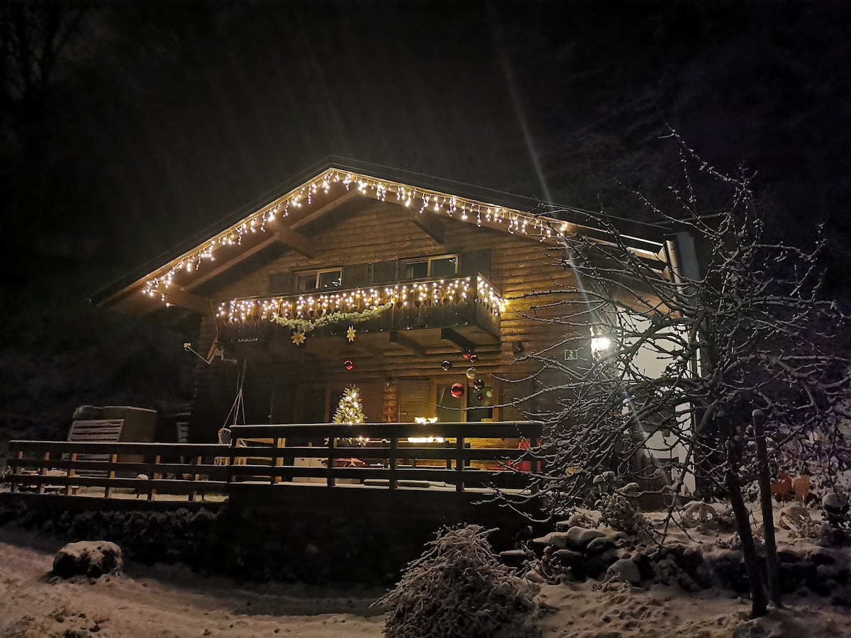 A charming wooden house is illuminated by warm string lights, casting a welcoming glow against the night sky. The snowy landscape surrounds the property, while a festive tree is visible on the porch. A wooden fence and trees contribute to the serene atmosphere.