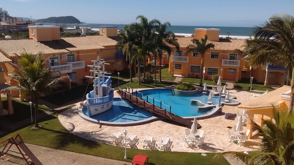 A bright outdoor area features a large pool surrounded by palm trees and lounge chairs. A playful ship-shaped play structure is positioned next to the pool, with a bridge leading across. The ocean can be seen in the background against a clear blue sky.