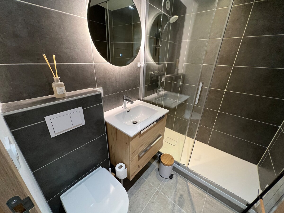 A modern bathroom features a spacious shower enclosed with clear glass. A circular mirror is mounted above a wooden vanity with a sink and storage. A toilet is positioned next to the vanity, and minimalistic décor is present, including a small stool and decorative items.