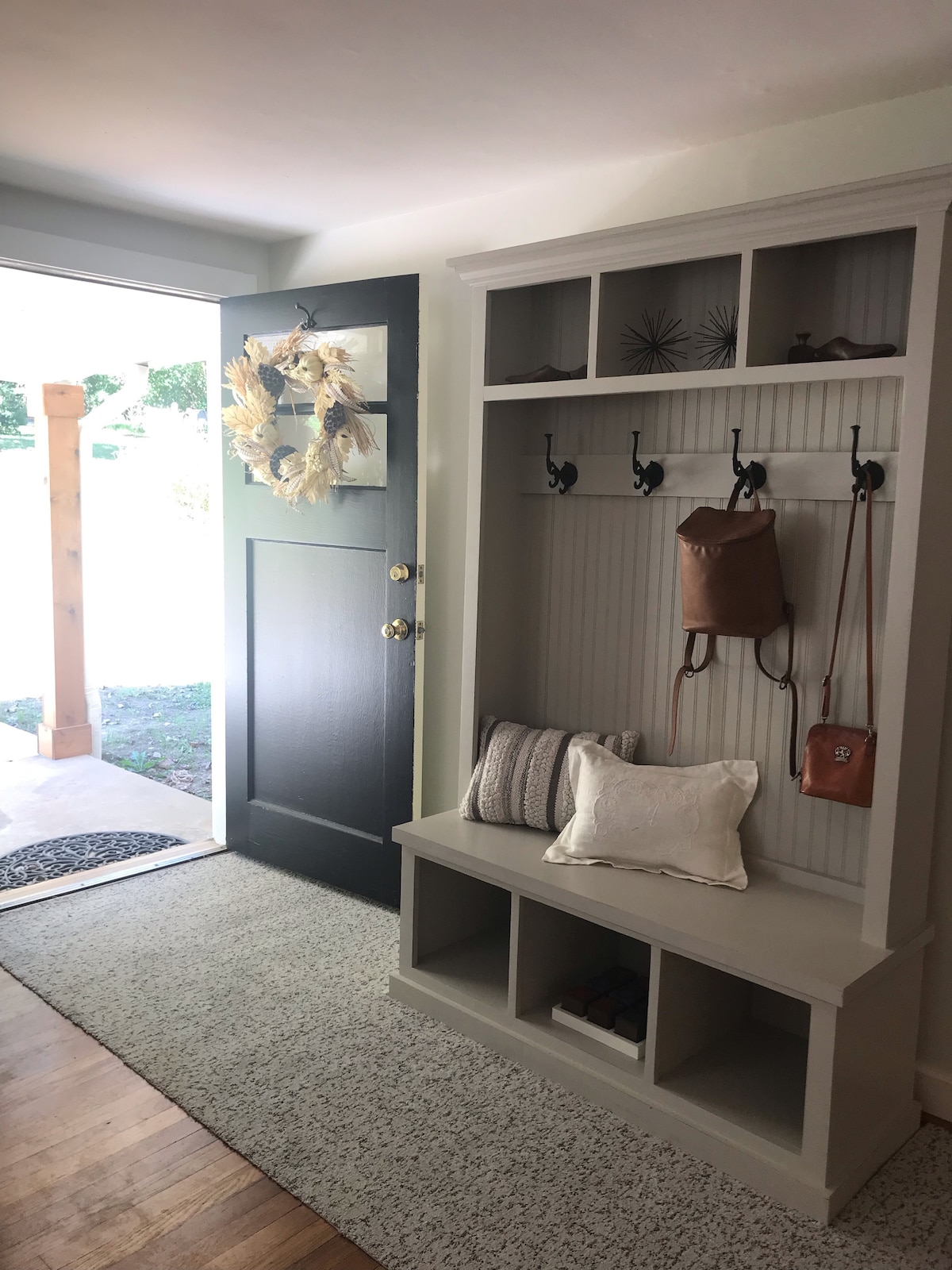 A welcoming entryway features a gray bench with cubby storage and hooks above for hanging bags and accessories. A door adorned with a wreath opens to a bright outdoor space, while a textured area rug adds comfort beneath the feet.