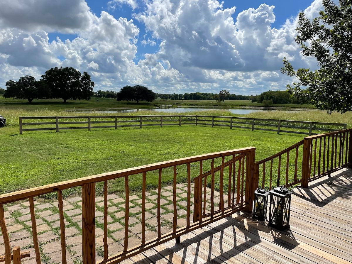 A spacious wooden deck offers views of a lush green yard, framed by a wooden fence. The landscape features open fields and distant trees, with a calm body of water visible. A few decorative lanterns sit near the edge of the deck.