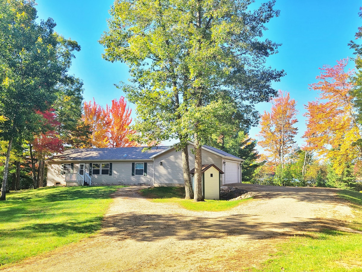 A spacious lake house is set on a large, well-maintained yard surrounded by vibrant autumn foliage. The exterior features a light-colored facade with large windows. A gravel driveway leads to the entrance, complementing the tranquil natural landscape.