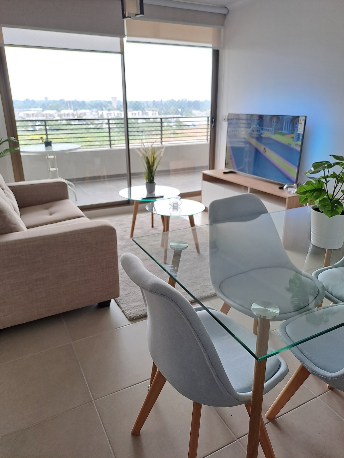 A modern living area is presented with a soft-colored sofa and glass tables. A flat-screen TV is mounted on the wall, and a plant adds a touch of greenery. Large windows offer a view of the surrounding area and allow natural light to fill the space.