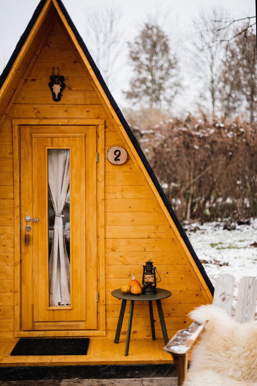An A-frame hut features a wooden exterior with a small porch area. The door is framed by a long window draped with sheer fabric. A designated number is displayed on the entrance. A small round table holds a lantern and decorative pumpkins beside a cozy chair.