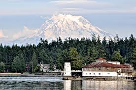 A majestic mountain is visible in the background, surrounded by lush evergreen trees. In the foreground, a calm body of water reflects the natural scenery. A small, distinctive structure is situated along the shoreline, highlighting the serene coastal environment.