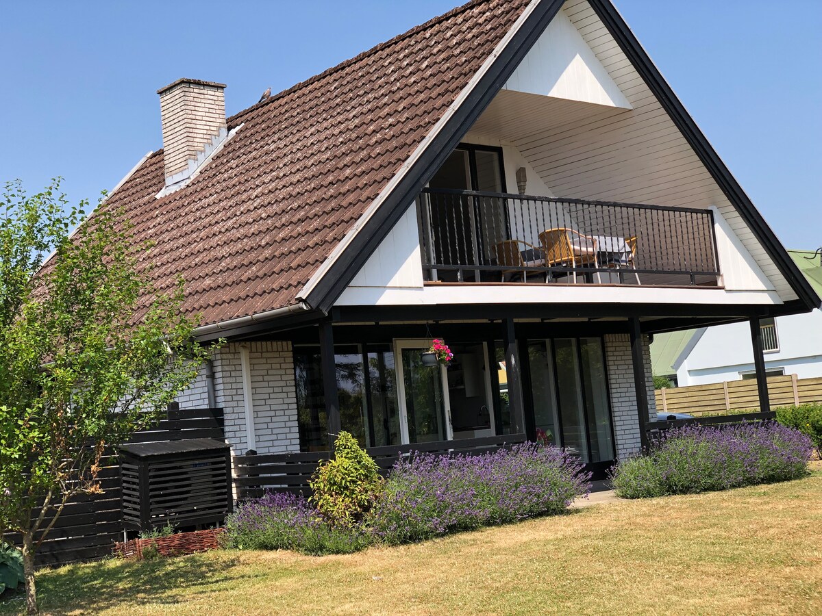 A two-story summer house is presented with a sloped roof and a balcony featuring outdoor seating. Lush greenery and blooming lavender are visible in the front yard. The exterior showcases a combination of light and dark tones, complemented by well-maintained grassy areas.