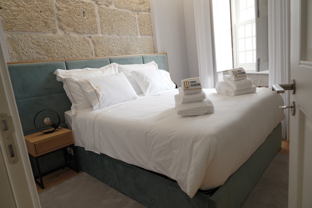 A cozy sleeping area features a spacious bed with crisp white linens, neatly arranged with two folded towels on each side. Light enters through a window adorned with white curtains, complementing the stone wall's texture.