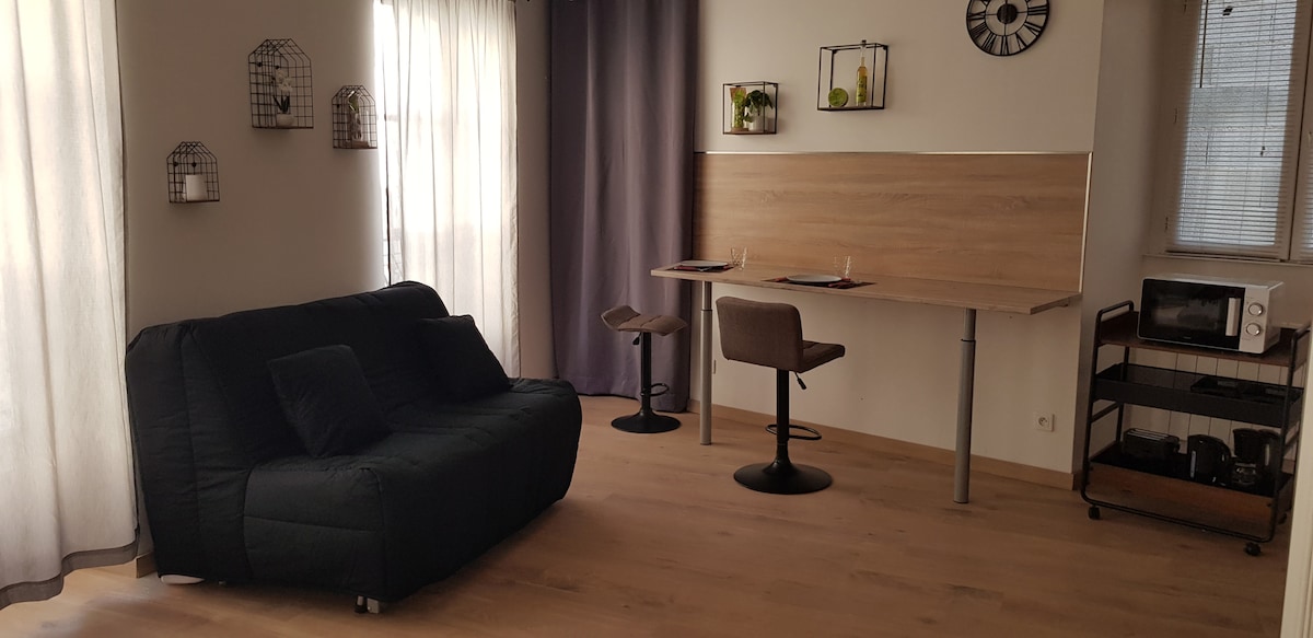 A living area features a dark sofa positioned adjacent to a wooden workstation. Two stools accompany the workspace, while a small cart with a microwave sits nearby. Natural light filters through the sheer curtains, enhancing the open and minimalistic design.