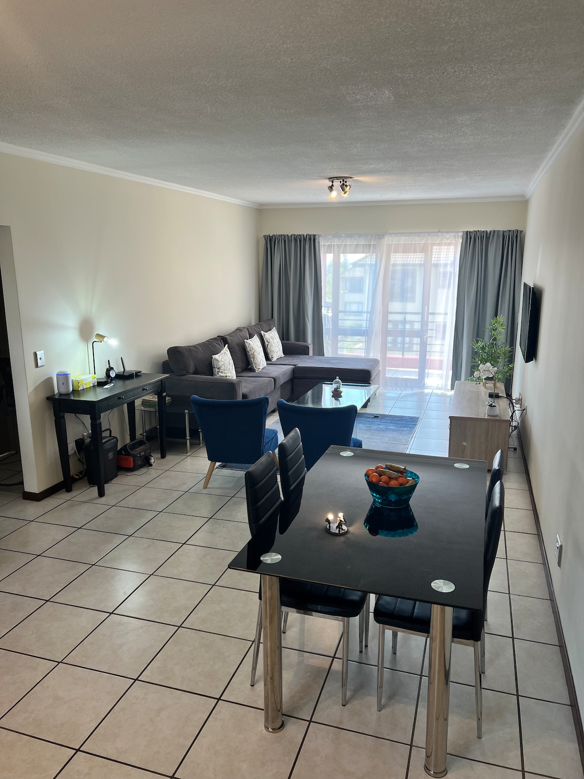 A spacious living area features a comfortable grey sofa, a dining table with four chairs, and a side table. Natural light enters through large glass doors, leading to a private balcony. Decorative elements include a bowl of fruit and soft textiles, creating a welcoming environment.