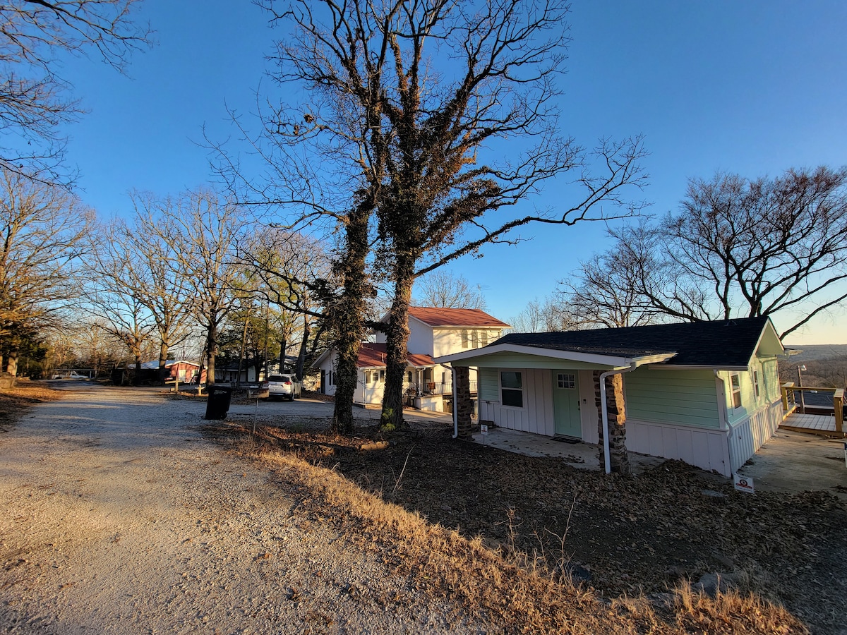 A tranquil view of the property shows a tree-lined pathway leading to the cabin. Several trees provide shade, and the rustic exterior of the cabin blends with the natural surroundings. The gravel driveway is visible, with additional homes in the background under a clear sky.
