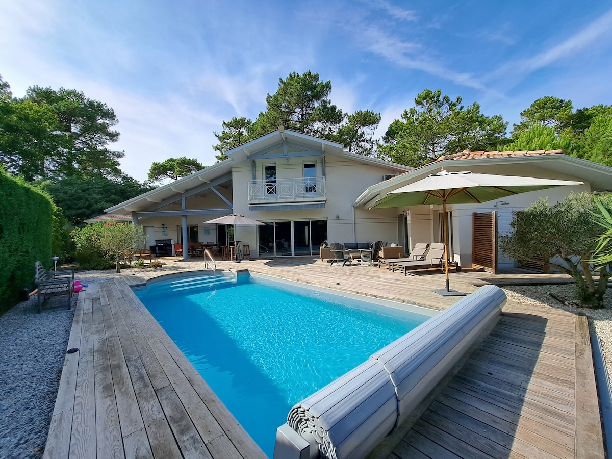 The outdoor area features a heated swimming pool surrounded by wooden decking. Lounge chairs and umbrellas provide shaded seating. The villa, characterized by a spacious terrace with glass doors, is nestled among trees, enhancing the serene ambiance of the garden.