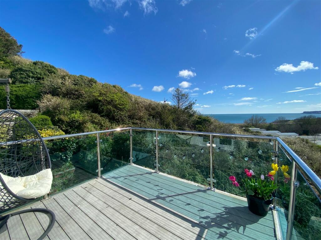 A glass balcony extends outward, offering unobstructed views of the sea and surrounding greenery. A wooden deck is present, adorned with a potted arrangement of colorful flowers. The clear blue sky complements the serene atmosphere, enhancing the space as a relaxing outdoor area.