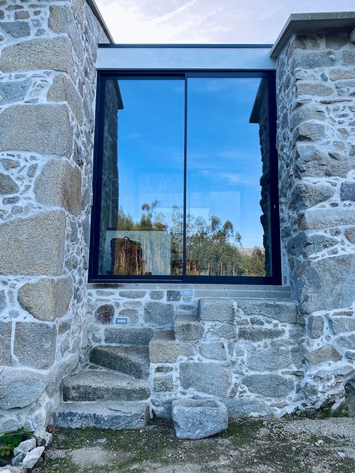 An exterior view of a stone wall structure is displayed, characterized by large glass panels reflecting the surrounding nature. The rugged texture of the stones contrasts with the sleek lines of the window. Steps lead up to the entrance, surrounded by a natural landscape.