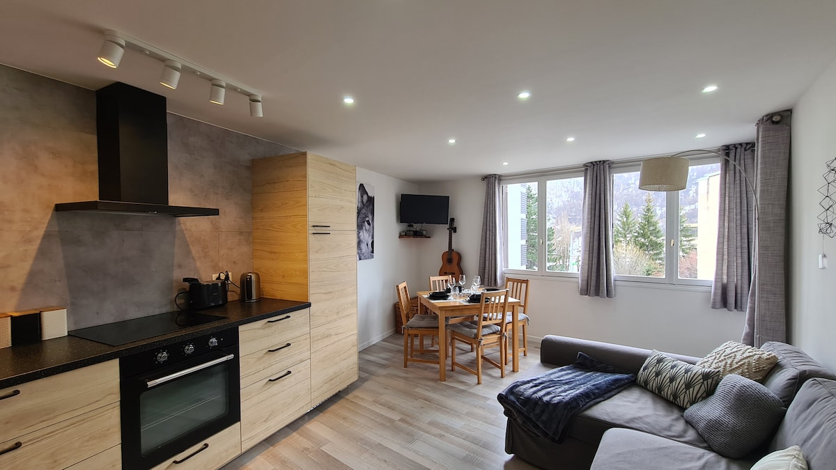 The open living area features a modern kitchen with wooden cabinetry and a sleek black countertop. A dining table with wooden chairs is visible, accompanied by a soft sofa adorned with a throw. Natural light enters through multiple windows, illuminating the space.