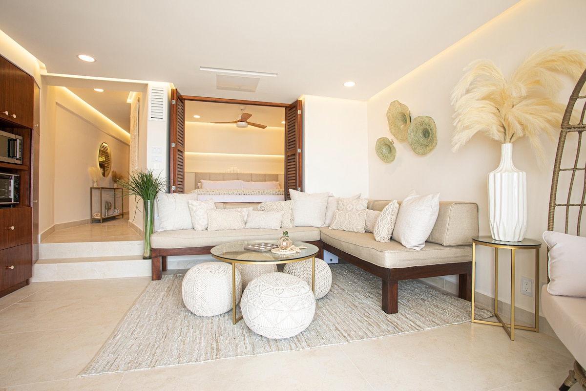 A spacious living room features a light-colored sectional sofa arranged around a round coffee table with soft decorative elements. Neutral tones are complemented by a cozy area rug, while decorative wall art and plants add subtle touches of detail in the modern design.