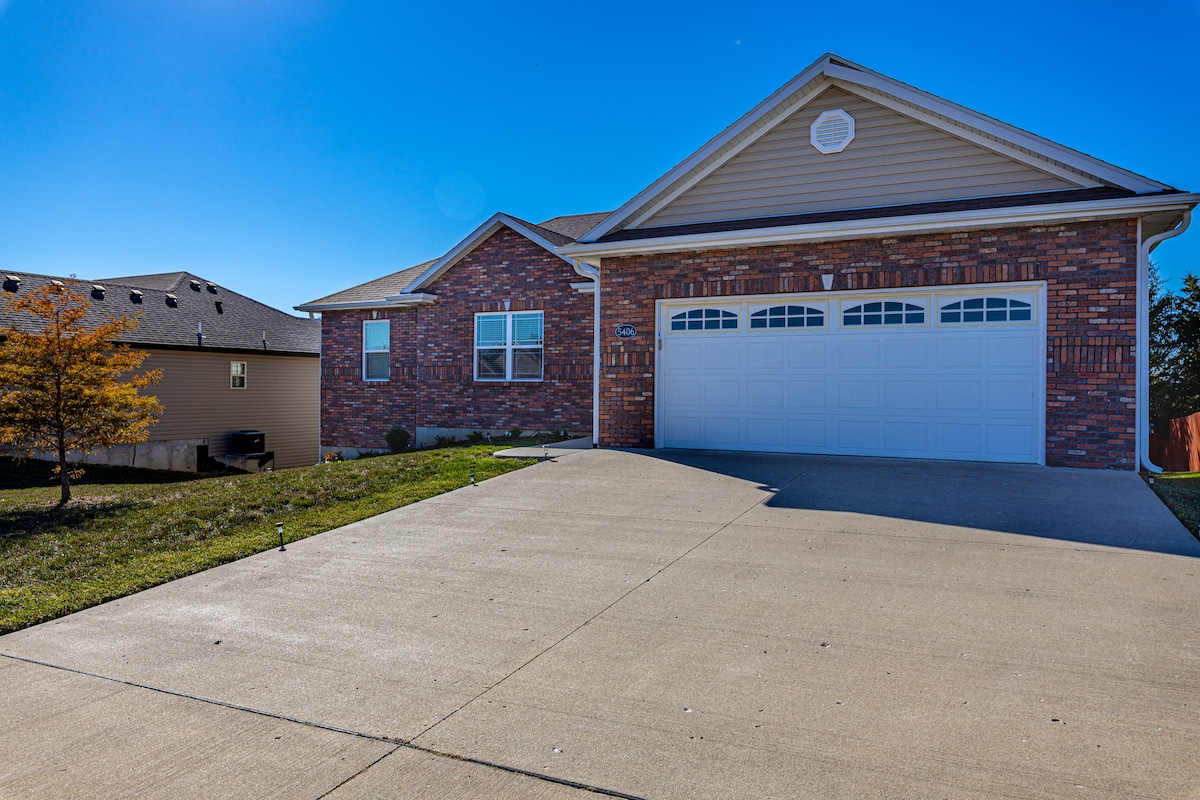A well-maintained exterior of a single-story home is shown, featuring a brick facade and light siding. The driveway leads to a two-car garage with decorative doors. A clear blue sky offers a bright backdrop, and landscaped areas are visible on either side of the driveway.