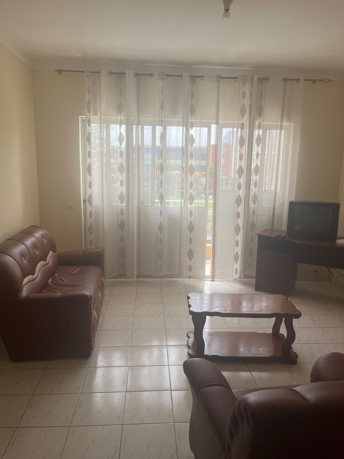 A living area is presented with two brown leather sofas and a wooden coffee table in the center. Natural light filters through sheer, patterned curtains, illuminating a small television on a wooden stand against the wall.