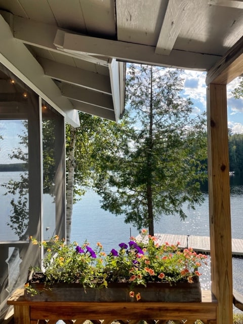A flower box filled with colorful blooms sits on the porch, framed by wooden railings. The tranquil water of Seymour Cove is visible in the background, with a wooden dock extending into the lake and trees providing a natural border.