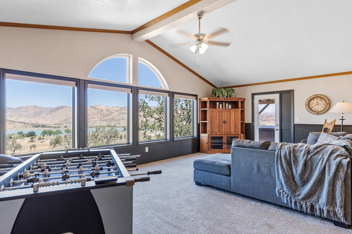The living area features a spacious sectional sofa draped with a cozy blanket, adjacent to a foosball table. Expansive windows frame stunning views of the valley and hills, while a ceiling fan enhances airflow. A wooden cabinet with a clock complements the natural light-filled space.