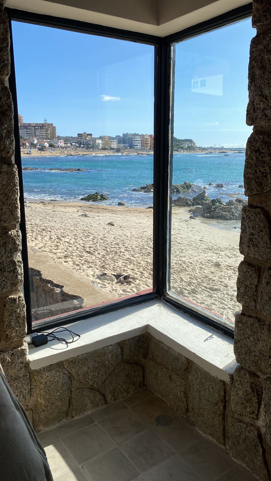 A serene view of the beach is framed by a large, clear window with stone accents. The sandy shore and rolling waves are visible, highlighting the proximity to the water. Sunlight brightens the scene, creating a peaceful atmosphere inside.
