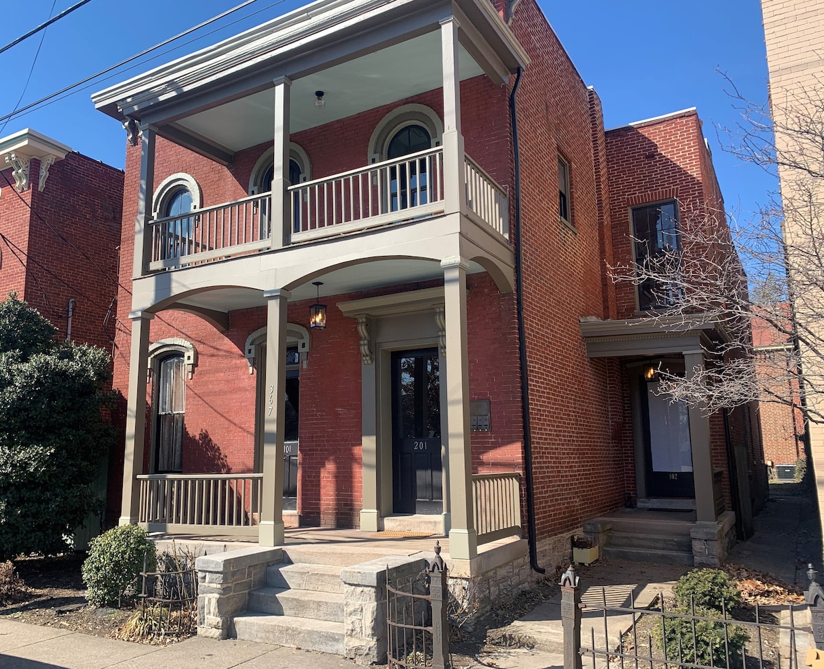 The exterior view captures a two-story brick home featuring a front porch supported by arched pillars. Balconies are visible on the second floor, and a well-maintained garden area frames the entrance. The design reflects the historical charm of the Italianate architectural style.