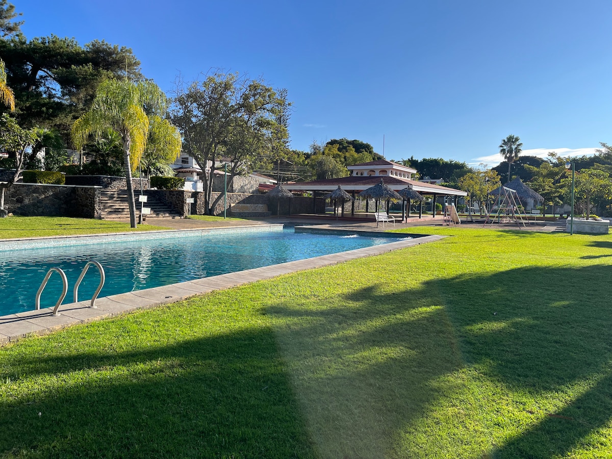 A spacious outdoor area features a sunlit swimming pool surrounded by lush green grass. Multiple shaded seating areas are present, along with recreational amenities visible in the background, including playground equipment and palm trees, creating an inviting space for relaxation and leisure.