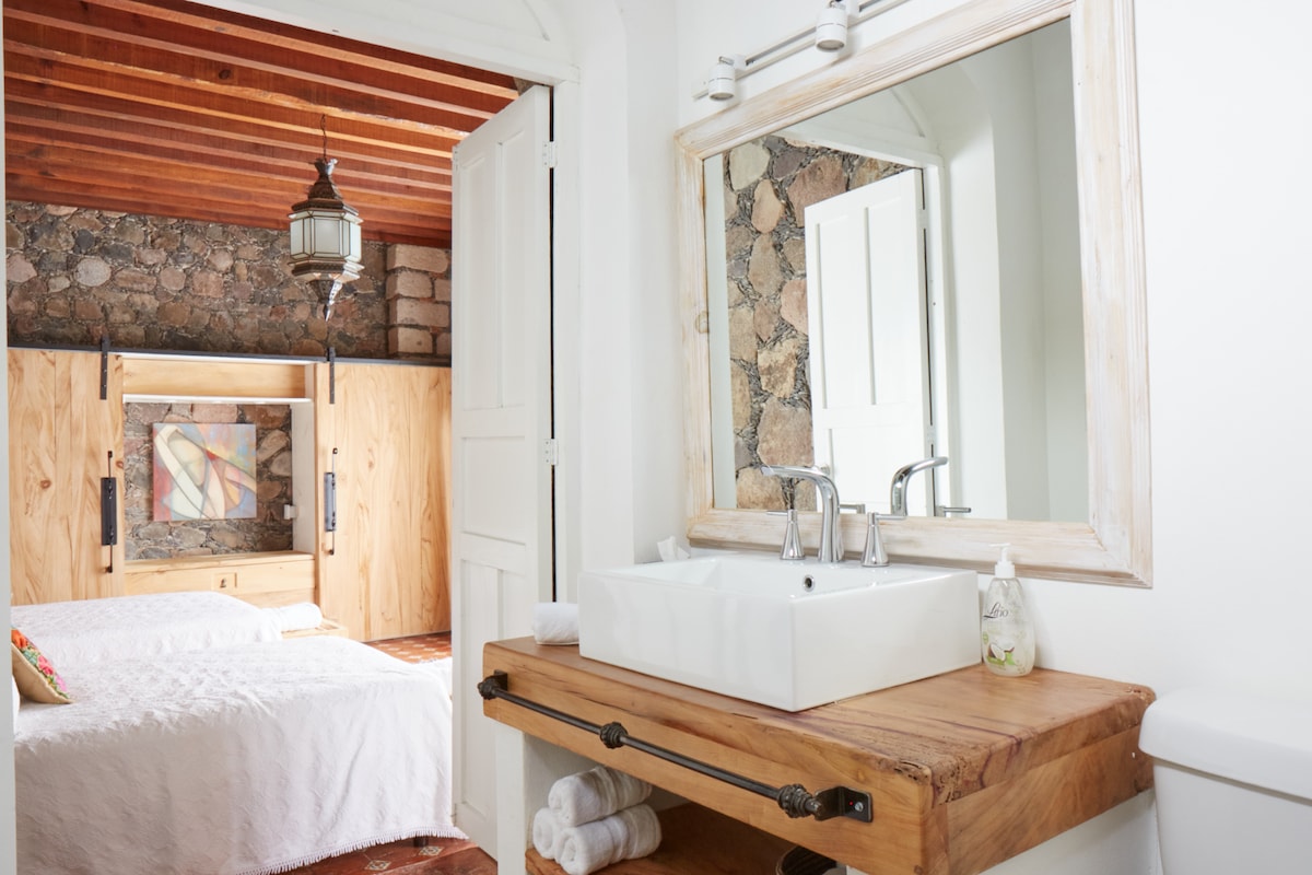A spacious bathroom area features a sleek white sink atop a wooden counter, enhanced by natural light from an adjacent window. The large mirror reflects elements of the tranquil bedroom, including two double beds and textured stone walls, creating a harmonious atmosphere.