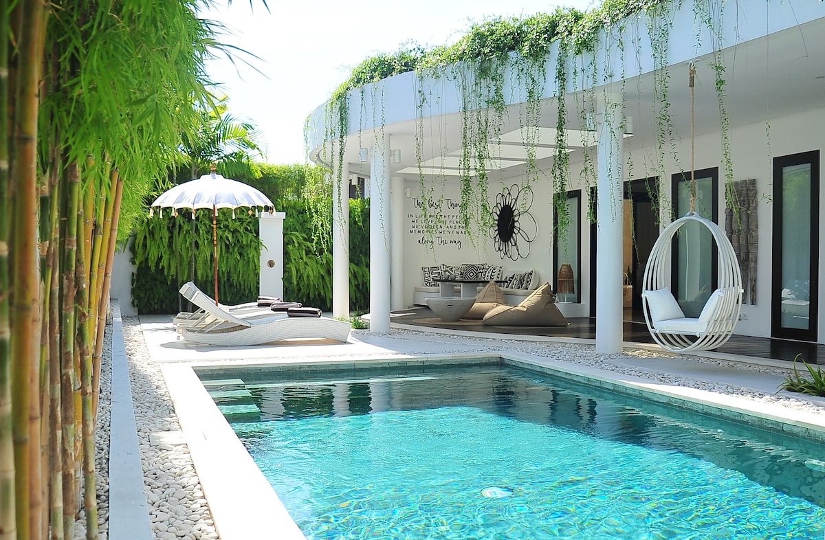 A serene private villa is depicted, featuring a clear swimming pool surrounded by tropical greenery and artistic decor. A hanging chair is visible next to a covered outdoor lounge area, emphasizing the inviting outdoor space. Sun loungers are arranged by the pool, enhancing the relaxation experience.
