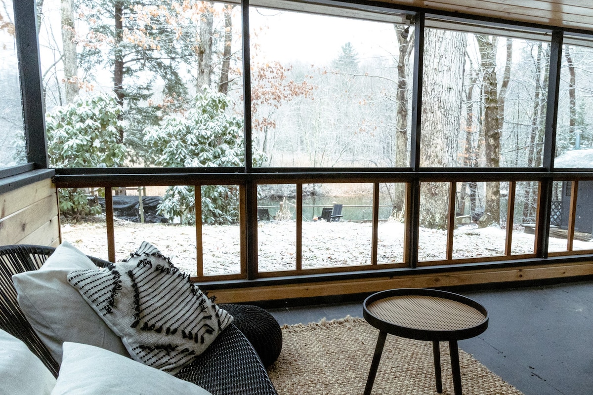 A screened-in porch offers a serene view of a winter landscape, featuring snow-dusted trees and a peaceful outdoor area. A small, round table is placed beside a lounge chair adorned with cushions, providing a cozy spot to relax indoors while enjoying the scenery.