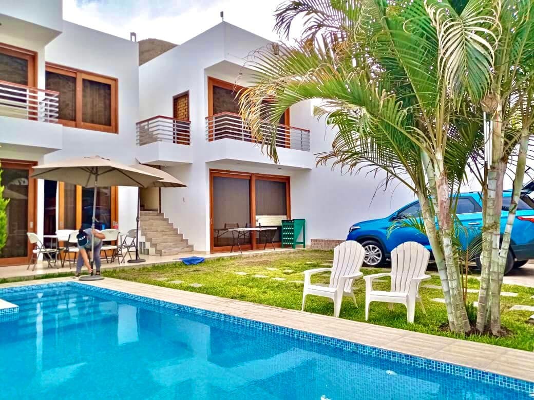 The outdoor area is showcased, featuring a clear blue pool surrounded by a manicured lawn. Two white lounge chairs provide a space for relaxation. A patio umbrella is positioned near a dining area, and a car is visible in the driveway.