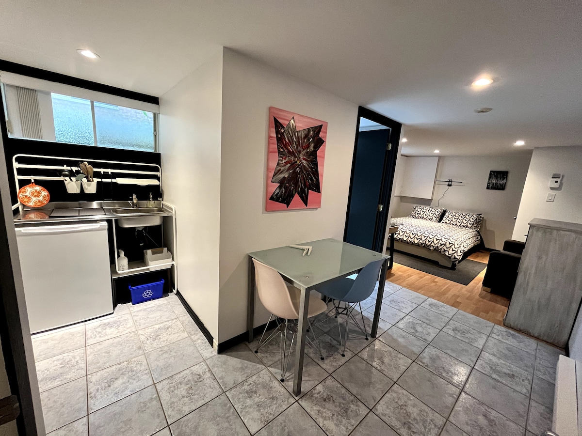 A modern studio space is revealed, featuring a small kitchenette with essential appliances and cookware. A glass dining table with two chairs is positioned in the center. A cozy sleeping area is visible with a patterned bedspread, complemented by wall art and natural light from a nearby window.