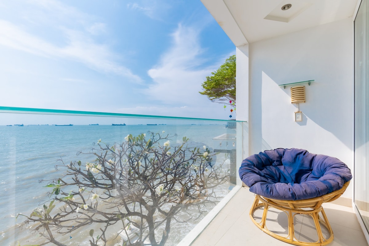 A private balcony overlooks a calm sea, with a comfortable blue cushioned chair positioned to enjoy the view. A flowering tree is visible nearby, and ships can be seen on the horizon under a clear sky.