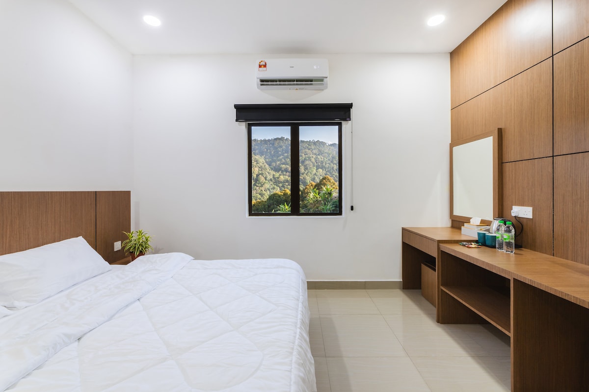 A spacious bedroom features a large bed with a minimalist white covering. A window offers a view of the surrounding mountains, while a wooden desk and mirror are positioned against the wall. Soft lighting illuminates the room, creating a calming environment.