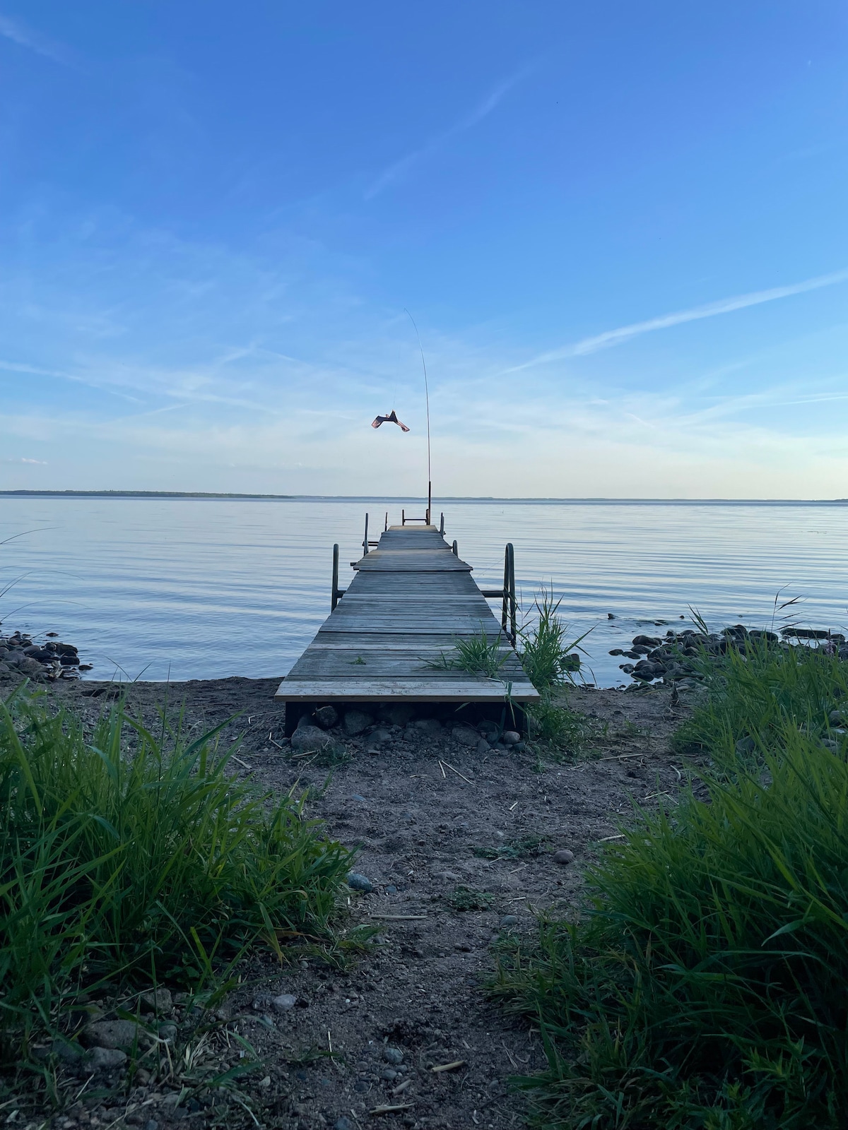 A wooden dock extends over a calm lake, flanked by gentle grass and stones. The water reflects a clear sky, with soft hues of blue and white. A lone figure can be seen in the distance, suggesting activity at the edge of the dock.