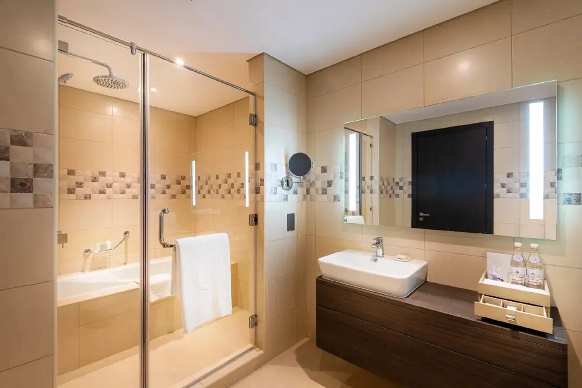 A modern bathroom is presented with a spacious walk-in shower, featuring beige tiled walls and integrated lighting. A sleek sink is positioned on a dark wooden vanity, while a large mirror reflects the clean lines. Bath towels are neatly arranged on a shelf.