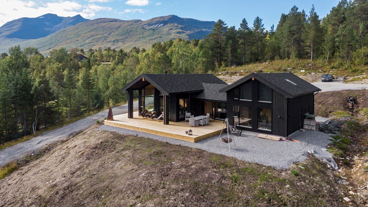 A modern cabin is nestled against a backdrop of rolling hills and trees, featuring a spacious wooden deck. Large windows maximize natural light and offer views of the surrounding landscape. A gravel path leads up to the cabin, enhancing its tranquil setting in nature.