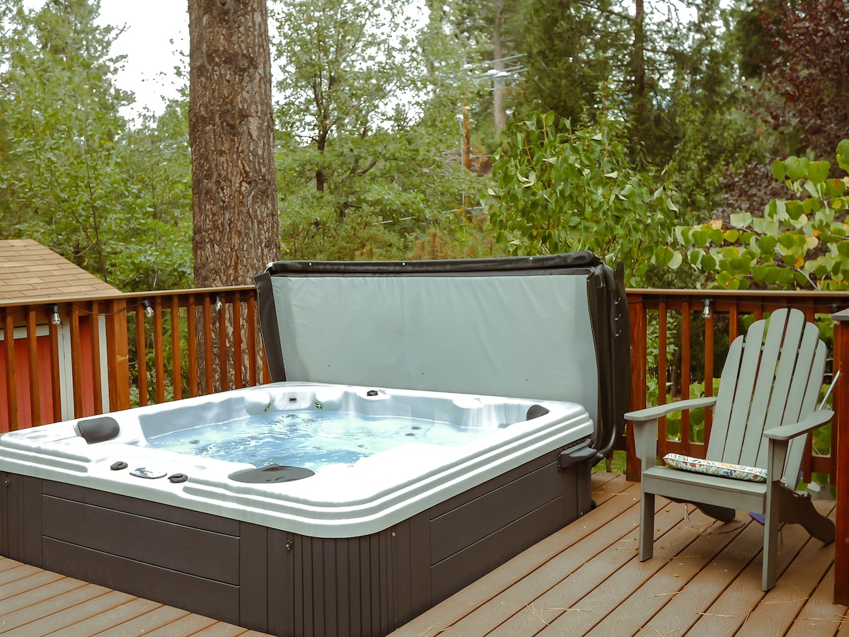 A hot tub is set on a wooden deck, surrounded by lush greenery and tall trees. A single adirondack chair is positioned nearby, offering a spot to relax. The hot tub cover is open, revealing the inviting water within.