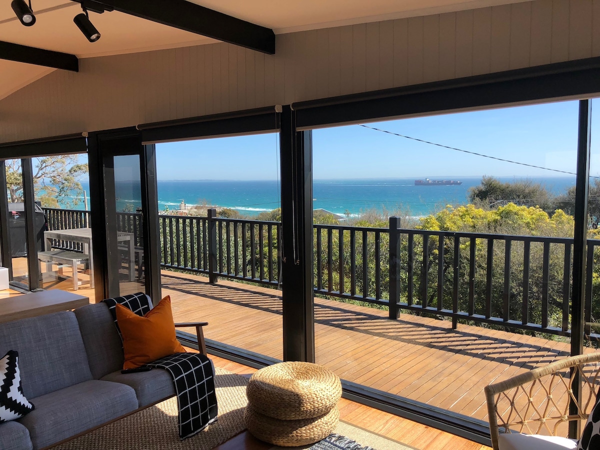 The living area features large glass sliding doors that open to a spacious deck with unobstructed views of the bay. Natural light fills the room, highlighting a cozy seating arrangement. The serene ocean scene is framed by lush greenery in the foreground.