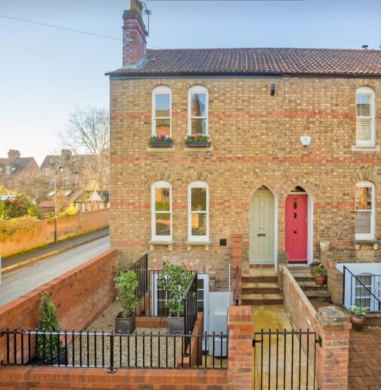 The exterior of a charming brick annex is showcased, featuring a beige front door and a bright red door, adding contrasting colors. The entrance is flanked by decorative planters, with a landscaped area visible in front, and the surrounding neighborhood includes trees and a paved walkway.
